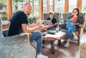 Mother, father and son family sitting together at Sunroom in cozy armchairs and reading books, using laptop or browsing a smartphone.